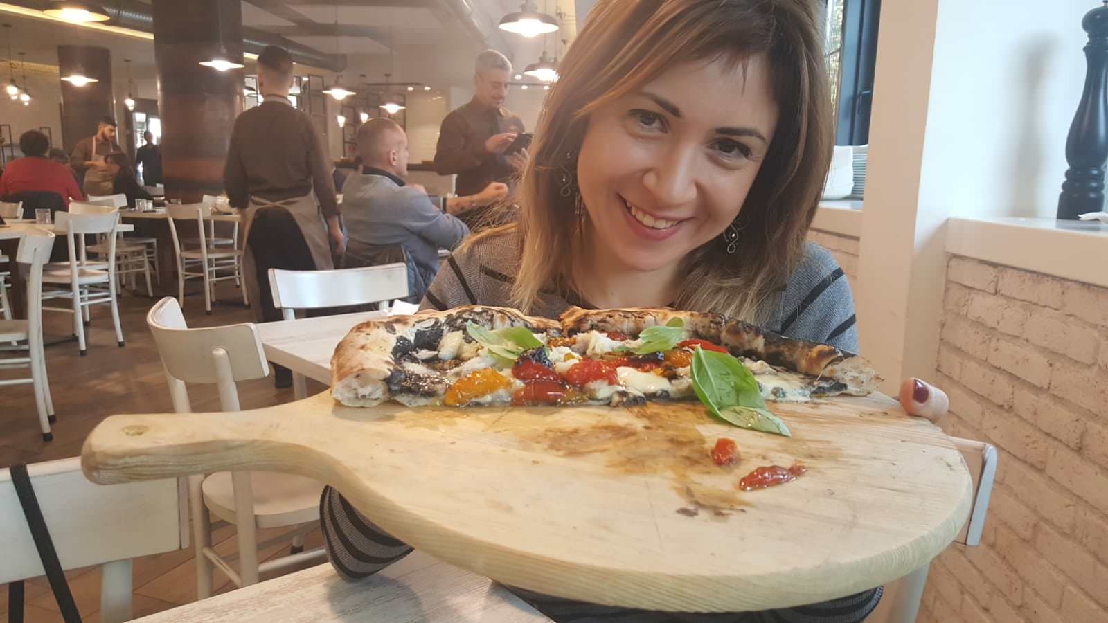 A woman is holding a wooden board towards the camera with a half eaten pizza on it and smiling